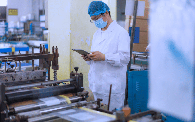 A lab technician in protective gear recording notes on a tablet in a manufacturing facility.